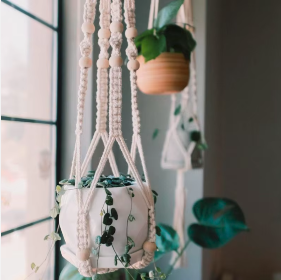 Porte-plante suspendu en macramé à perles de bois, pot blanc et plante retombante devant une fenêtre