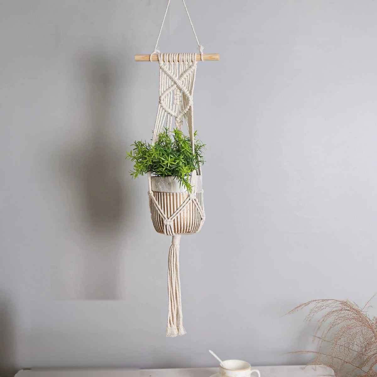 Suspension pot de fleurs macramé - bâton de bois, pot clair et plante verte devant un mur gris